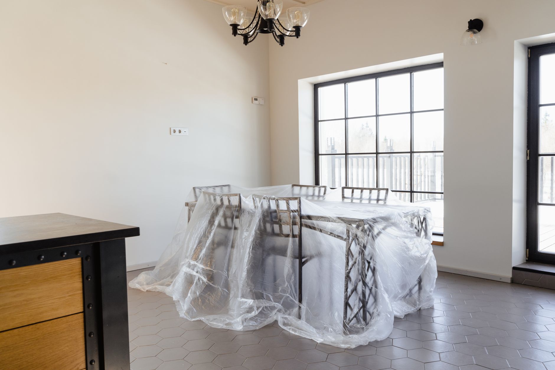 Dining table and chairs wrapped in plastic sheeting beside a large black-framed window
